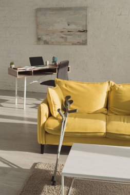 Sunlit living room with crutches near yellow sofa  clipart