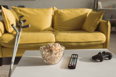 KYIV, UKRAINE - JANUARY 21, 2020: Selective focus of popcorn, remote controller and joystick with crutches near couch in living room