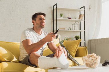 KYIV, UKRAINE - JANUARY 21, 2020: Selective focus of man with broken leg watching tv near popcorn and joystick on coffee table