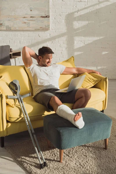 Sorrindo Homem Com Perna Quebrada Otomano Olhando Para Laptop Sofá — Fotografia de Stock