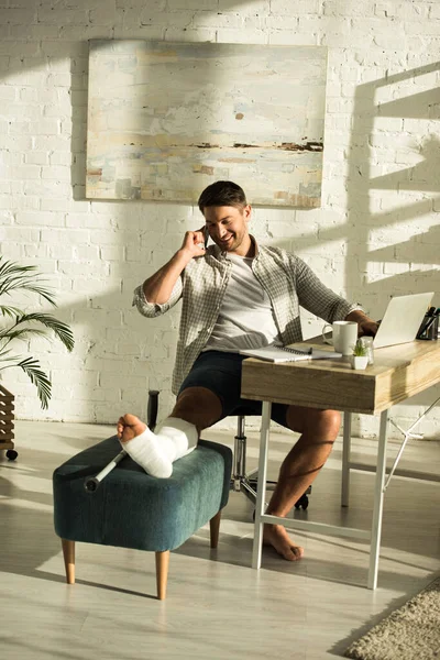 Smiling Freelancer Broken Leg Talking Smartphone While Sitting Table Laptop — Stock Photo, Image