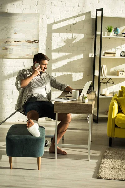Freelancer Con Pierna Rota Usando Portátil Hablando Smartphone Mesa Sala — Foto de Stock