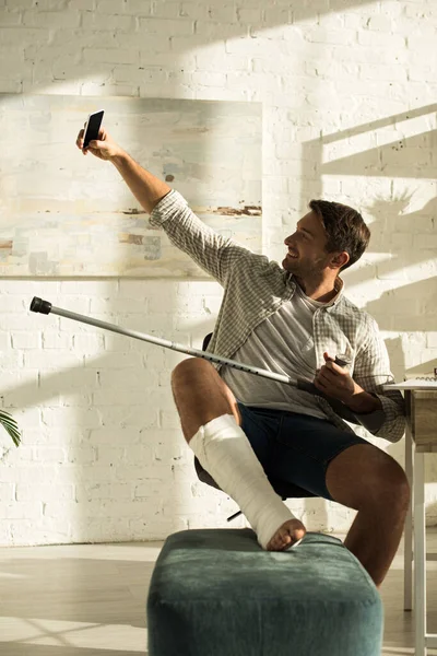 Smiling man with broken leg and crutch taking selfie with smartphone at home