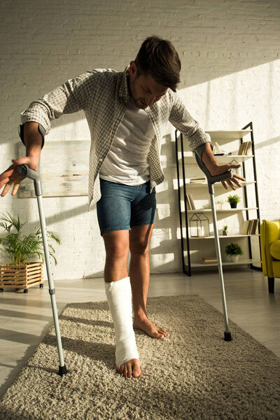 Красивый мужчина со сломанной ногой, держа костыли, стоя в гостиной
