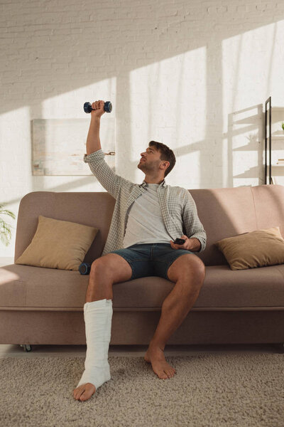 Handsome man with leg in plaster bandage training with dumbbell and holding remote controller on couch