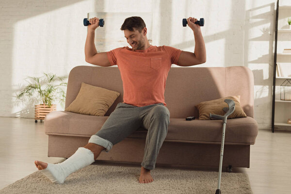 Smiling man with broken leg exercising with dumbbells on couch in living room