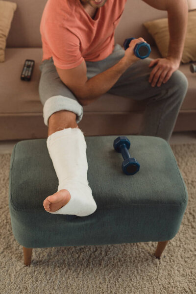 Cropped view of man with broken leg on ottoman working out with dumbbell on couch