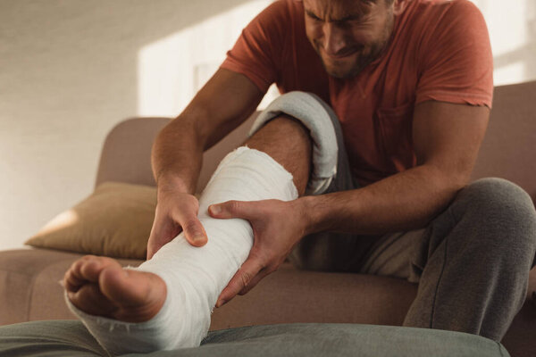 Man having pain while touching broken leg in plaster on couch