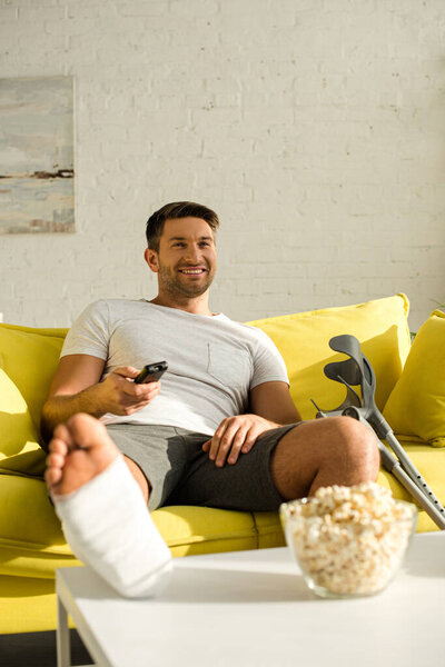 Selective focus of smiling man with leg in plaster bandage watching tv near popcorn on coffee table
