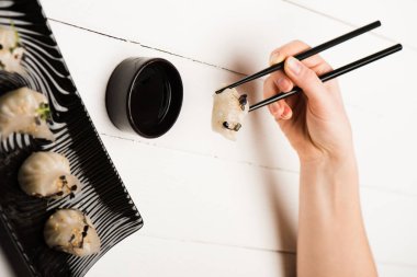 cropped view of woman eating delicious Chinese boiled dumplings with chopsticks and soy sauce at white wooden table