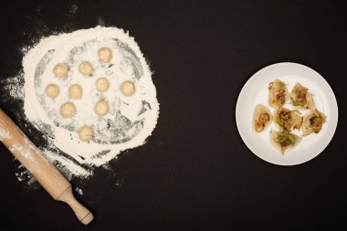 top view of delicious varenyky with cracklings on plate near raw dough balls, flour and rolling pin on black background