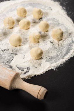 raw dough balls for dumplings on flour near rolling pin on black background
