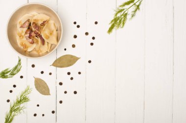 top view of delicious varenyky with cracklings and dill in bowl near bay leaves and black peppercorns on white wooden table