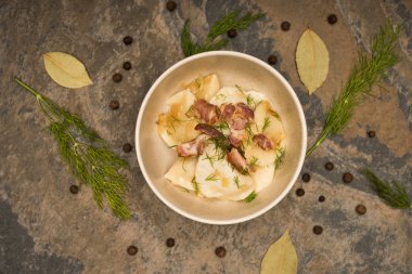 top view of delicious varenyky with cracklings and dill in bowl near bay leaves and black peppercorns on stone surface