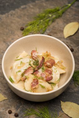 selective focus of delicious varenyky with cracklings and dill in bowl near bay leaves and black peppercorns on stone surface