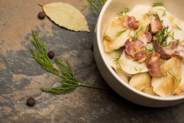delicious varenyky with cracklings and dill in bowl near bay leaves and black peppercorns on stone surface
