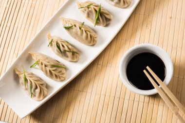 top view of delicious Chinese boiled dumplings on plate near chopsticks and soy sauce on bamboo mat