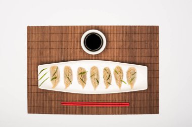 top view of delicious Chinese boiled dumplings on plate near chopsticks and soy sauce on bamboo mat on white background