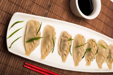 delicious Chinese boiled dumplings on plate near chopsticks and soy sauce on bamboo mat
