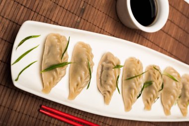 delicious Chinese boiled dumplings on plate near chopsticks and soy sauce on bamboo mat clipart