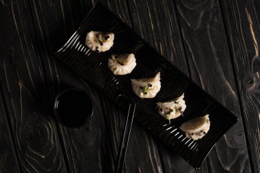 top view of delicious Chinese boiled dumplings on plate near chopsticks and soy sauce on black wooden table