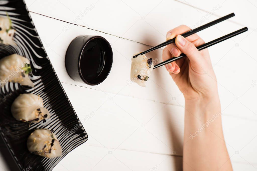 cropped view of woman eating delicious Chinese boiled dumplings with chopsticks and soy sauce at white wooden table