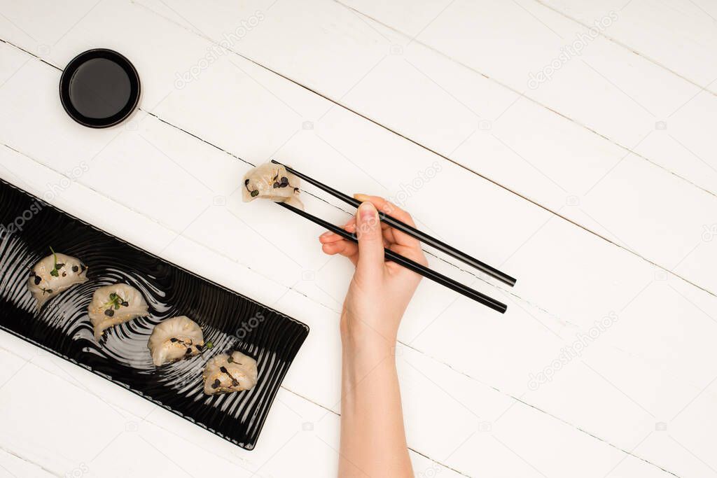 cropped view of woman eating delicious Chinese boiled dumplings with chopsticks and soy sauce at white wooden table