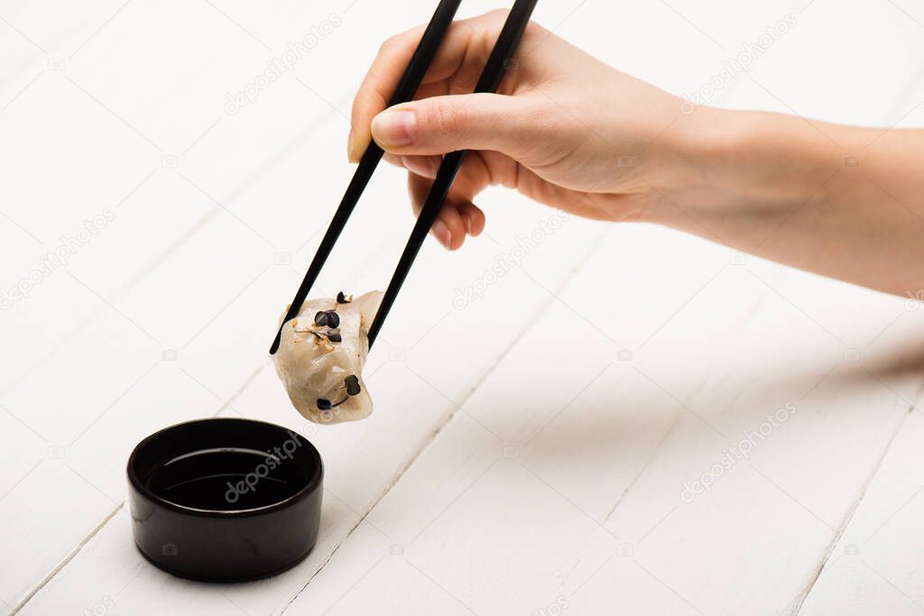 cropped view of woman eating delicious Chinese boiled dumpling with chopsticks and soy sauce at white wooden table