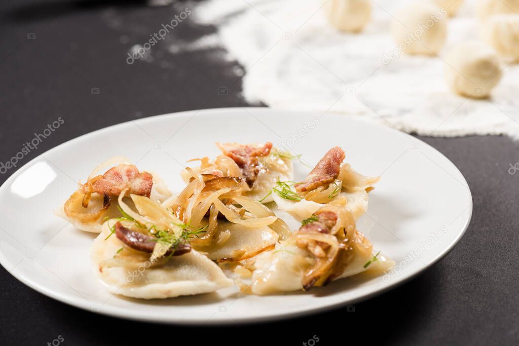 selective focus of delicious varenyky with cracklings on plate near raw dough balls, flour on black background