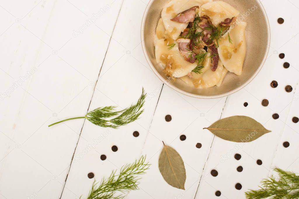 top view of delicious varenyky with cracklings and dill in bowl near bay leaves and black peppercorns on white wooden table