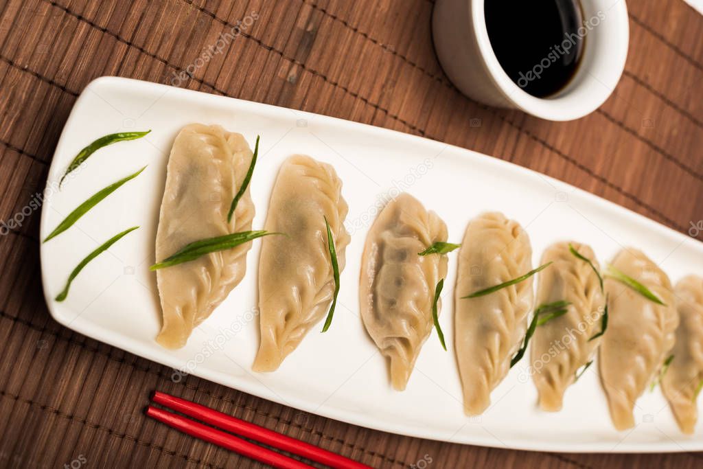 delicious Chinese boiled dumplings on plate near chopsticks and soy sauce on bamboo mat
