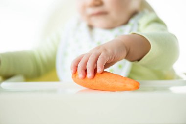 Selective focus of baby holding carrot while sitting on feeding char isolated on white