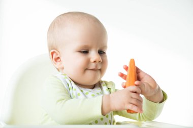 Adorable baby holding carrot while sitting on feeding chair isolated on white