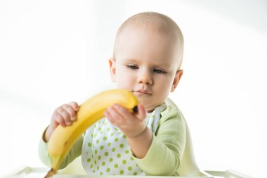 Selective focus of adorable baby holding ripe banana while sitting on feeding chair on white background