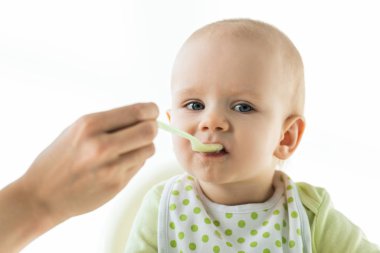 Selective focus of mother with spoon feeding baby with puree on feeding chair isolated on white clipart