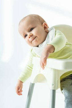 Cute baby with soiled mouth in puree sitting on feeding chair on white background
