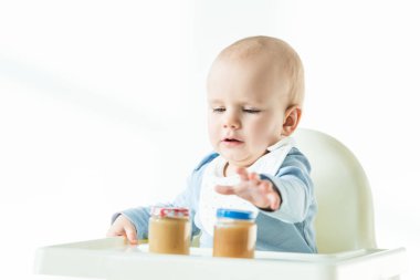 Selective focus of baby sitting on feeding chair with jars on fruit puree on table on white background