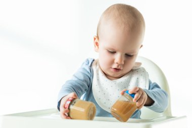 Baby holding jars of fruit puree while sitting on feeding chair on white background