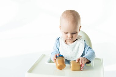 Adorable baby boy holding jars with baby food on table of feeding chair on white background