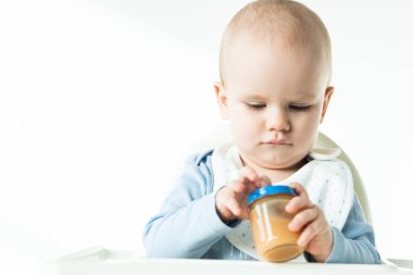 Selective focus of baby boy on feeding chair holding jar with fruit puree on white background
