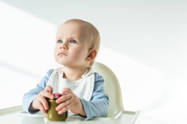 Selective focus of cute baby holding jar of vegetable baby food and looking away on feeding chair on white background