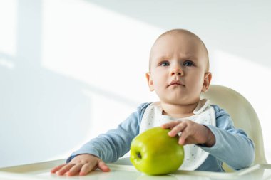 Cute baby boy touching green apple on table of feeding chair on white background