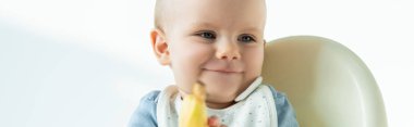 Selective focus of smiling baby boy holding banana on feeding chair on white background, panoramic shot
