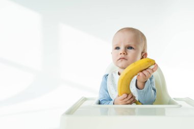 Baby boy holding ripe banana while sitting on feeding chair on white background