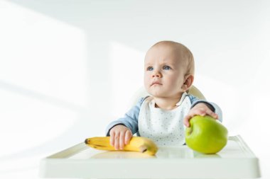 Adorable baby holding ripe banana and apple on table of feeding chair on white background