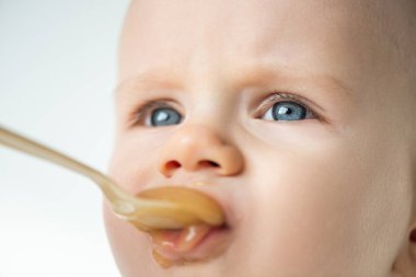 Close up view of adorable baby boy looking away during feeding with fruit puree isolated on grey 