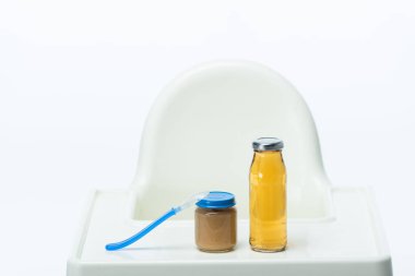 Jar of baby food with spoon and bottle of apple juice on table of feeding chair isolated on white