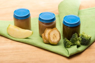 Jars of baby food with pieces of apple, broccoli and banana slices on napkin on wooden surface