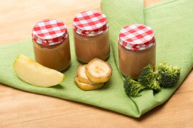 Jars of prepared baby nutrition with pieces of apple, broccoli and banana slices on napkin on wooden background