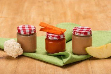 Pieces of apple, cauliflower and carrot near jars of baby food on napkin on wooden surface